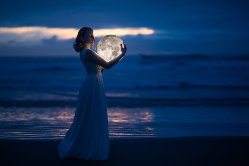 A woman in a long dress, standing by the ocean holding the moon at twilight; image is meant to depict a witch working magick with the moon