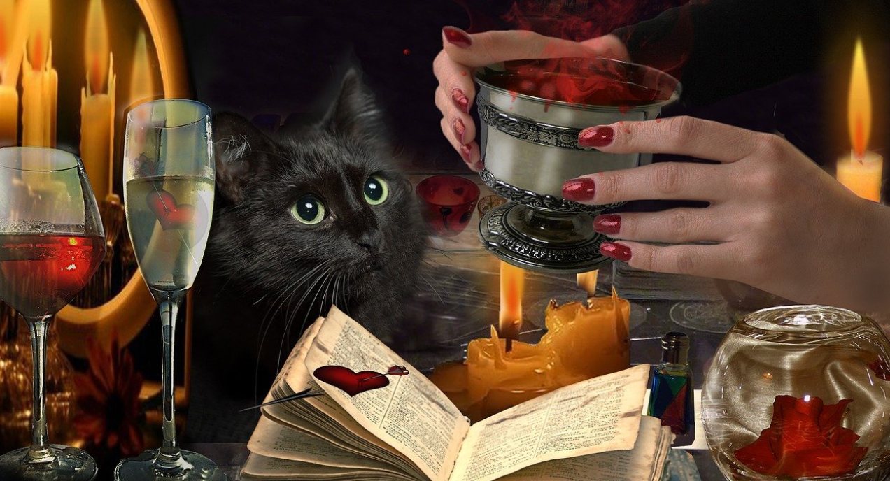 Full color image of a woman's hands with red fingernails holding a pewter goblet filled with a red magic potion that is smoking, alluding to it being a magick spell. It is assumed to be a love spell because there is an open book with a red blood heart on it. A black cat watches the spellcasting.